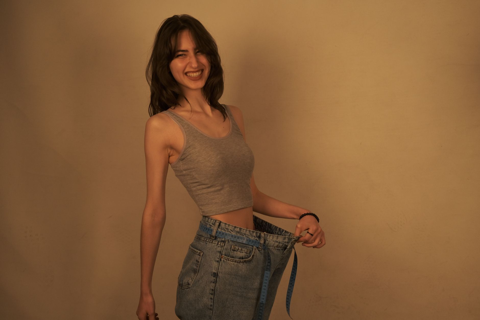 Smiling woman showcasing weight loss by wearing oversized jeans indoors.