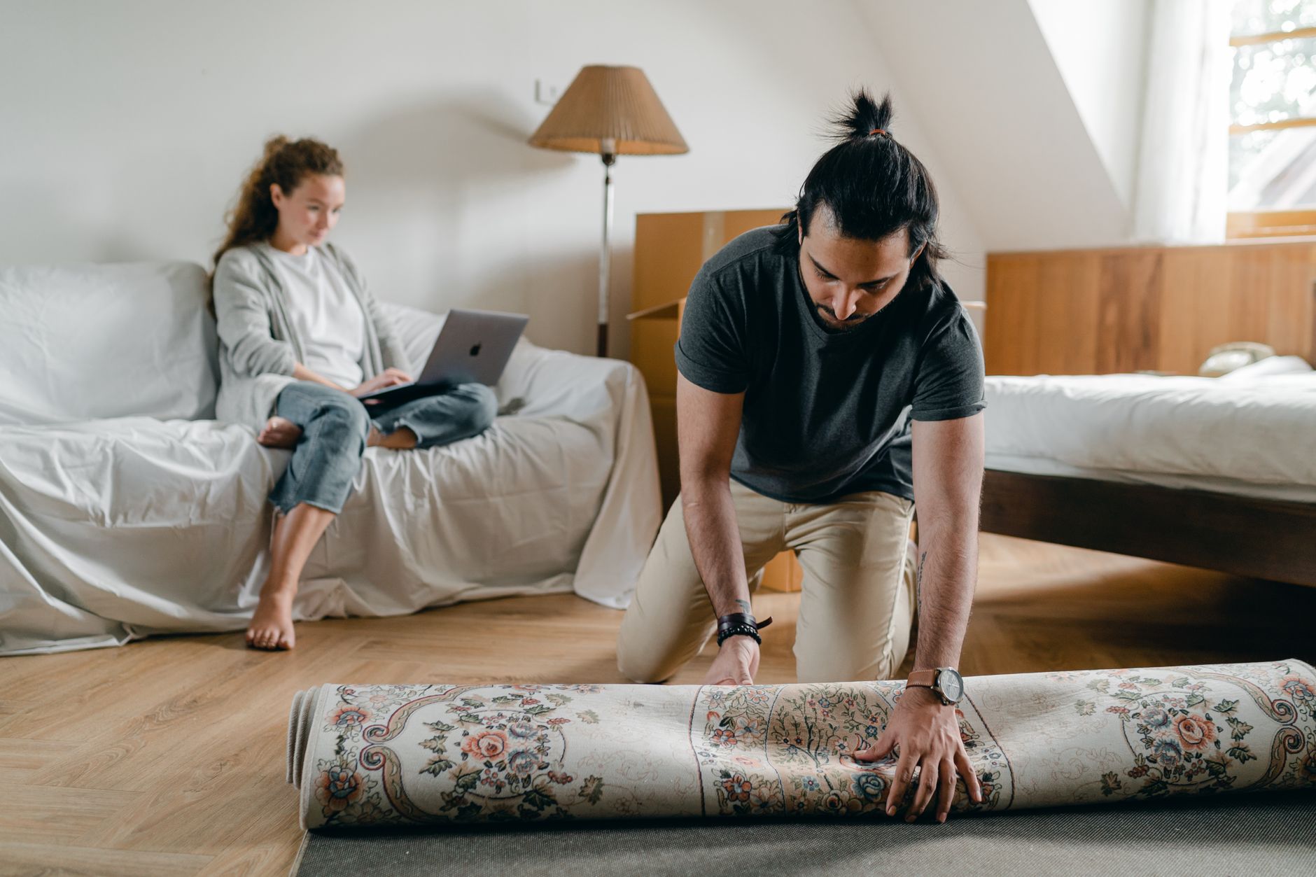 Fit ethnic male in wristwatch laying carpet on parquet while barefoot woman browsing internet on portable computer sitting on cozy sofa in bedroom