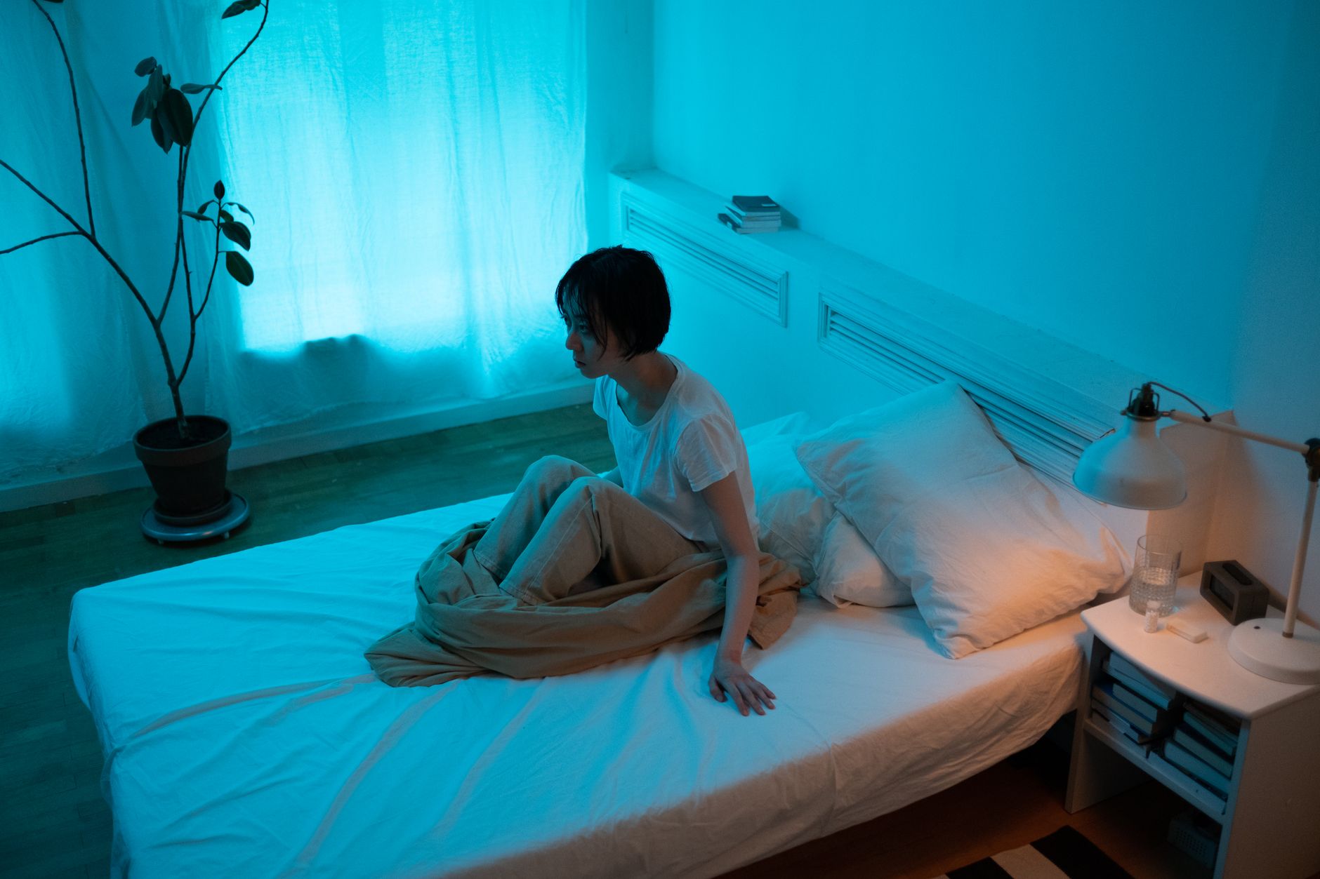 Woman sitting on bed in dimly lit room, conveying themes of insomnia and contemplation.