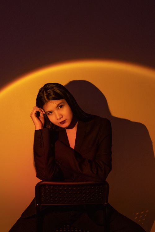 Free A woman in a black blazer poses creatively in warm studio lighting, casting a captivating shadow. Stock Photo