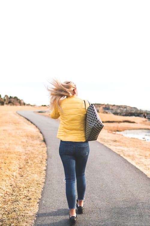 女性は灰色のアスファルトの道を歩く黄色のバブルジャケットを着ています