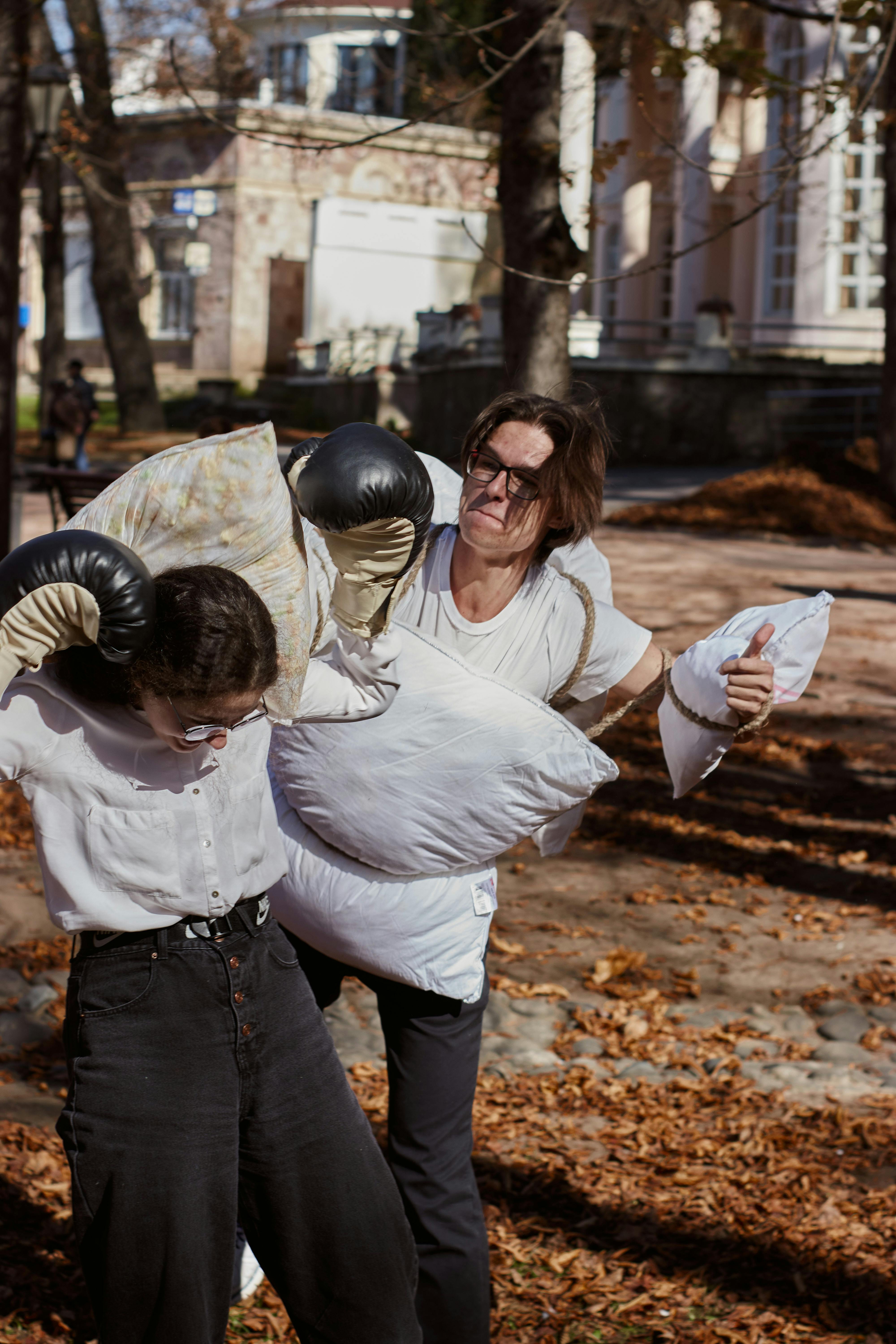 Two adults enjoy a playful pillow fight in an autumn park, surrounded by fallen leaves.