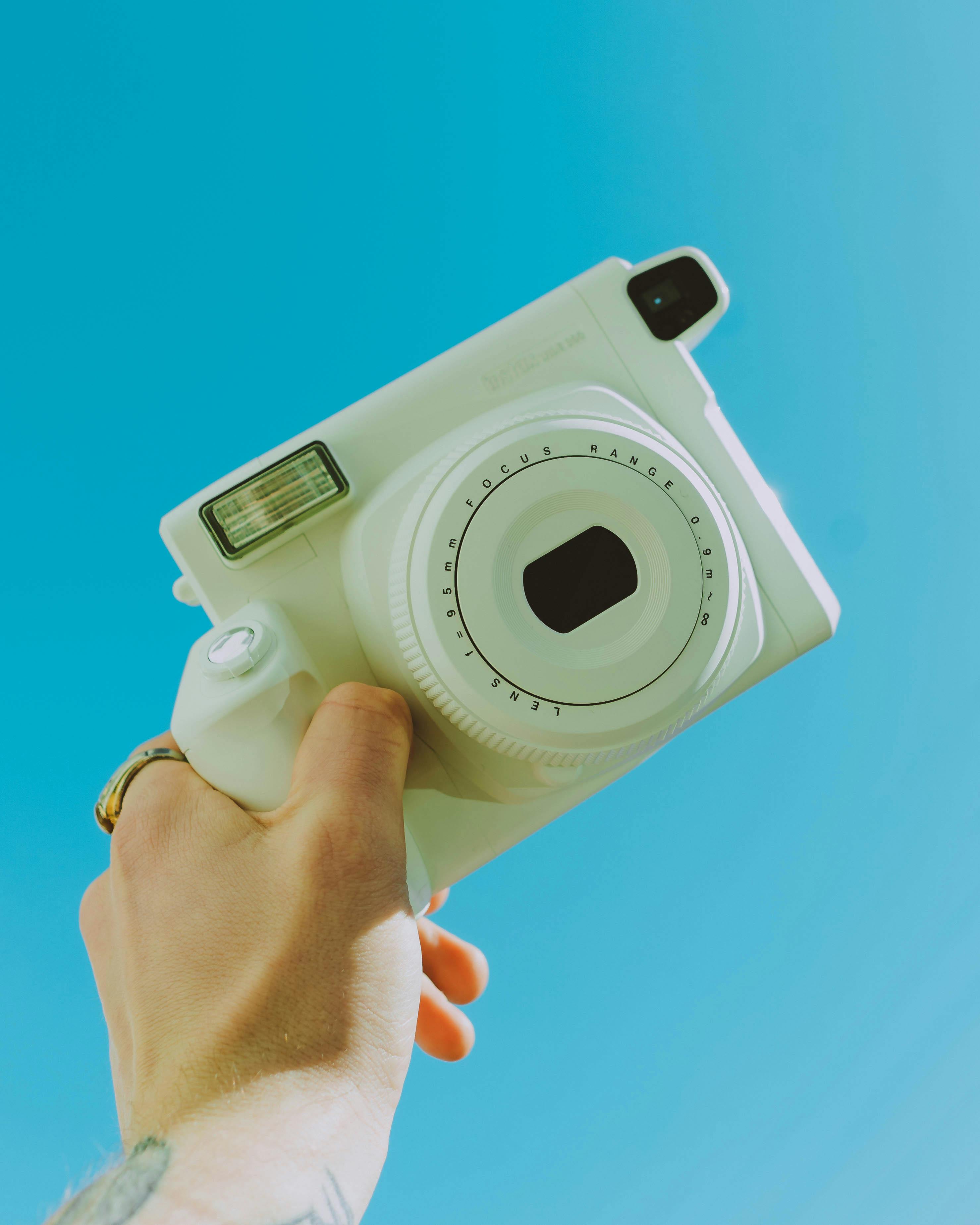 A hand holds a white instant camera up against a vibrant blue sky, capturing the essence of outdoor photography.
