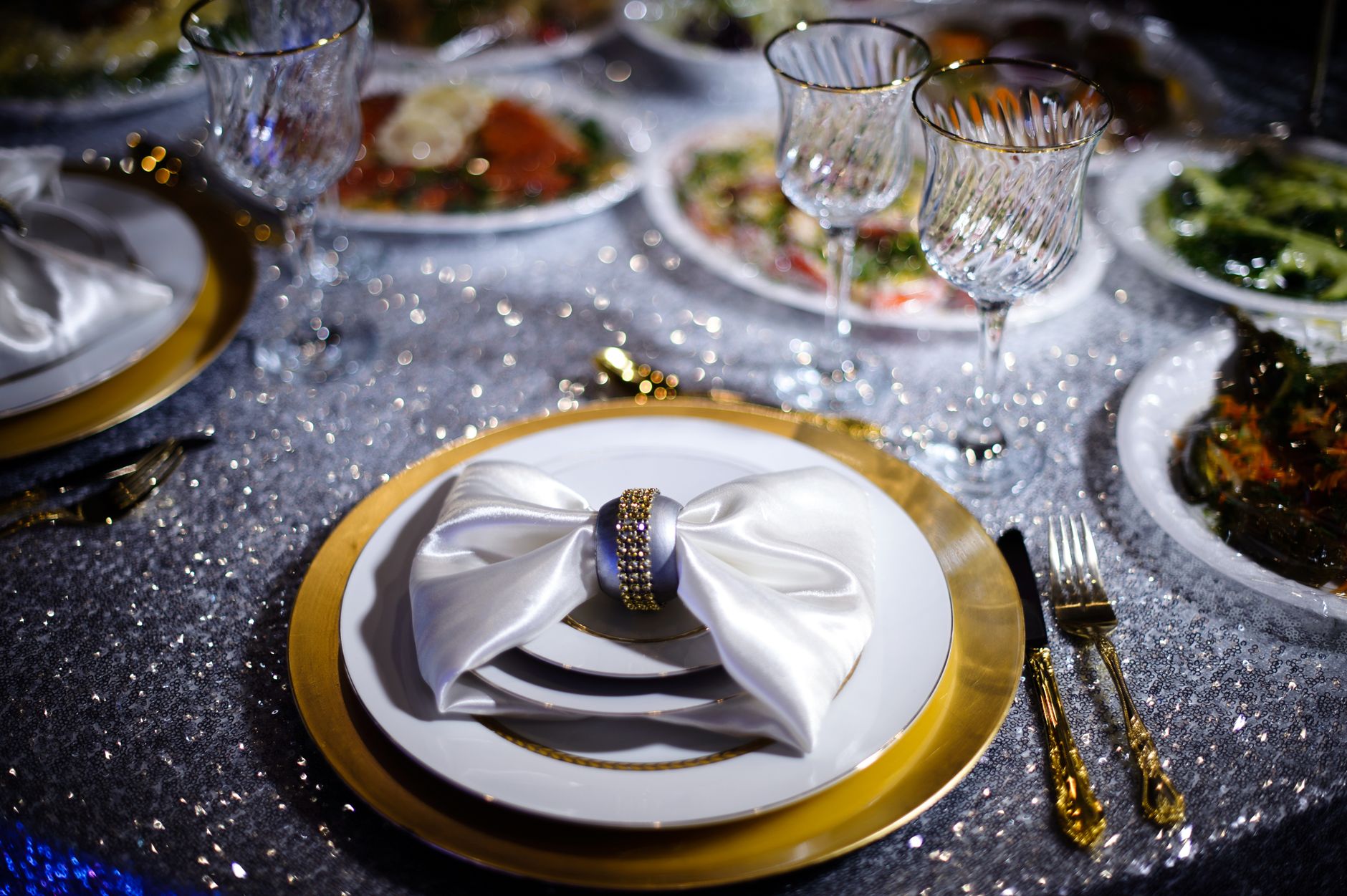 Luxurious table setup with gold cutlery, crystal glasses, and fine dinnerware on sparkling cloth.