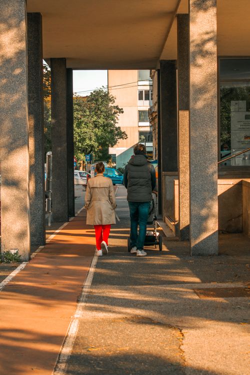 Free A couple walks with a stroller along a city sidewalk under trees, creating a warm urban scene. Stock Photo