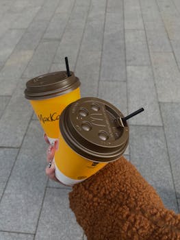 Close-up of a hand holding two takeaway coffee cups on a street.