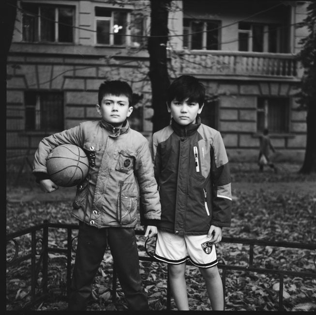 Two boys wearing jackets stand with a basketball in a courtyard, surrounded by buildings.