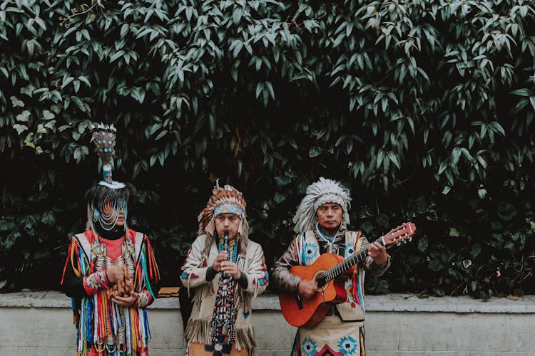 Three Men In Traditional Native American Clothes Playing Guitar On Side Of Road