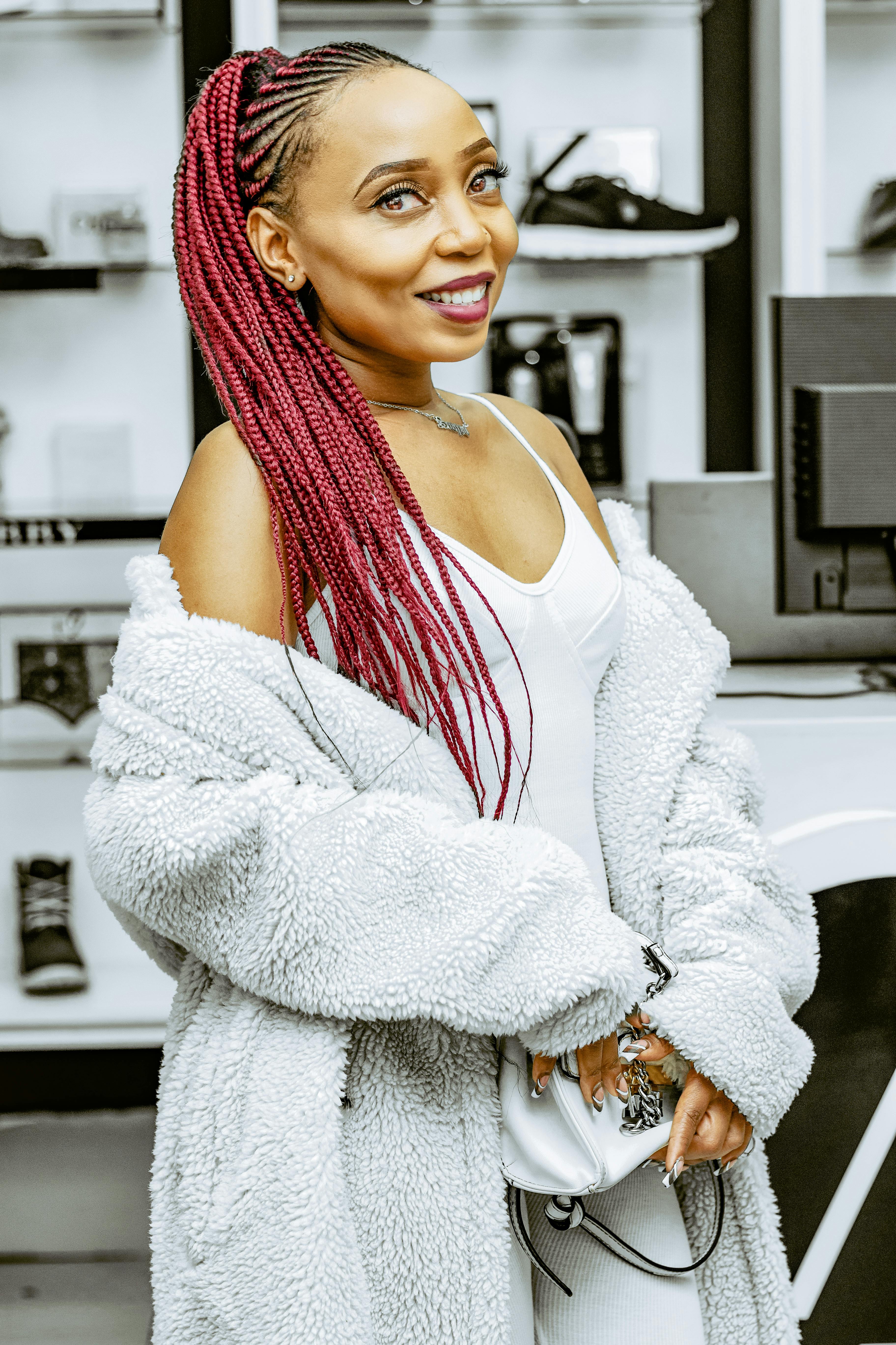 Confident woman with red braided hair wearing a white outfit and fluffy coat in a modern indoor setting.