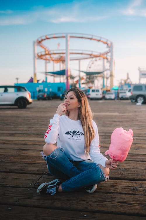 A Woman in White Sweater and Denim Jeans Sitting on a Wooden Floor while Holding a Cotton Candy