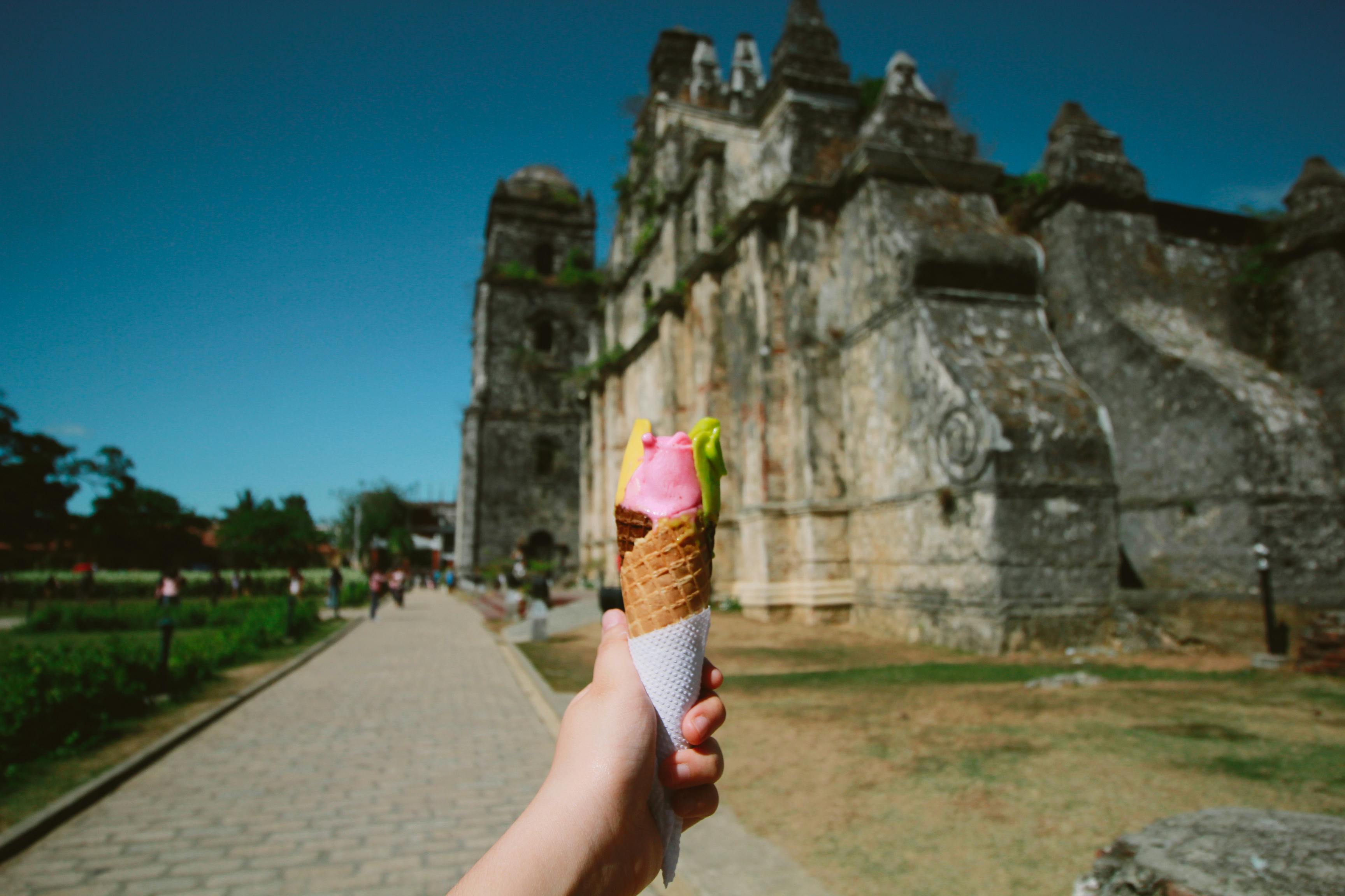 Hand holding vibrant ice cream cone near historic stone ruin under clear blue sky.