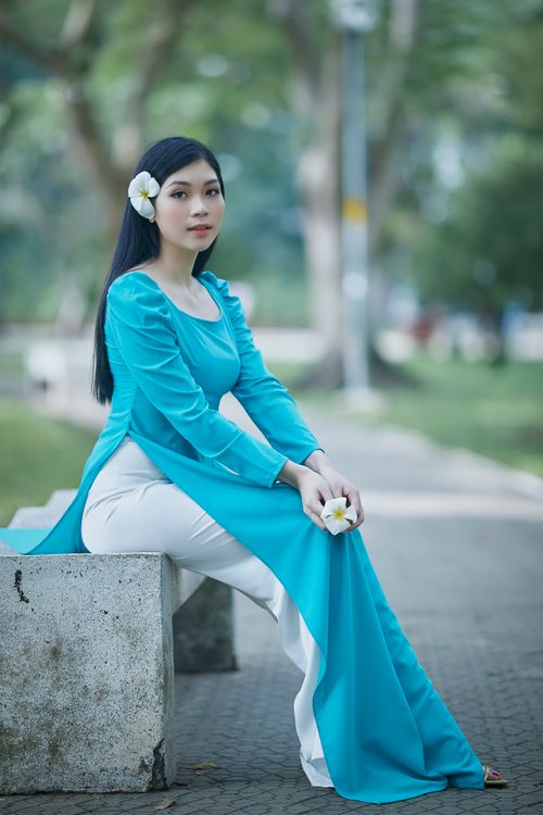 Free A woman in a blue dress with a plumeria flower sits on a bench in a lush park. Stock Photo