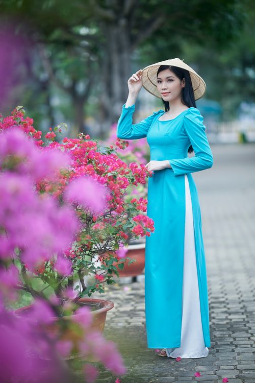 Free Asian woman wearing a blue dress and conical hat stands gracefully in a park with flowers. Stock Photo