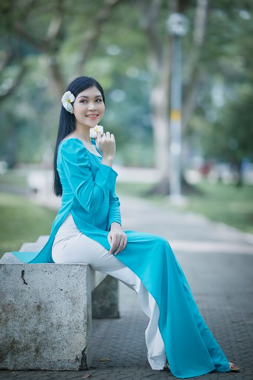 Free A woman wearing a blue Ao Dai sits gracefully on a park bench with flowers, exuding elegance. Stock Photo