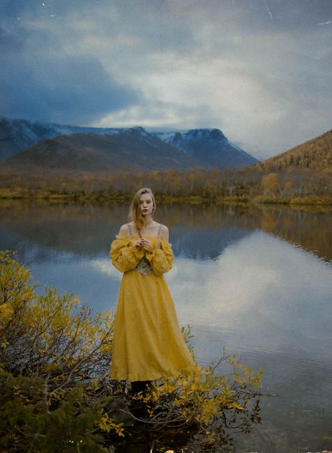 A woman in a yellow dress stands by a tranquil lake with mountain views during autumn.