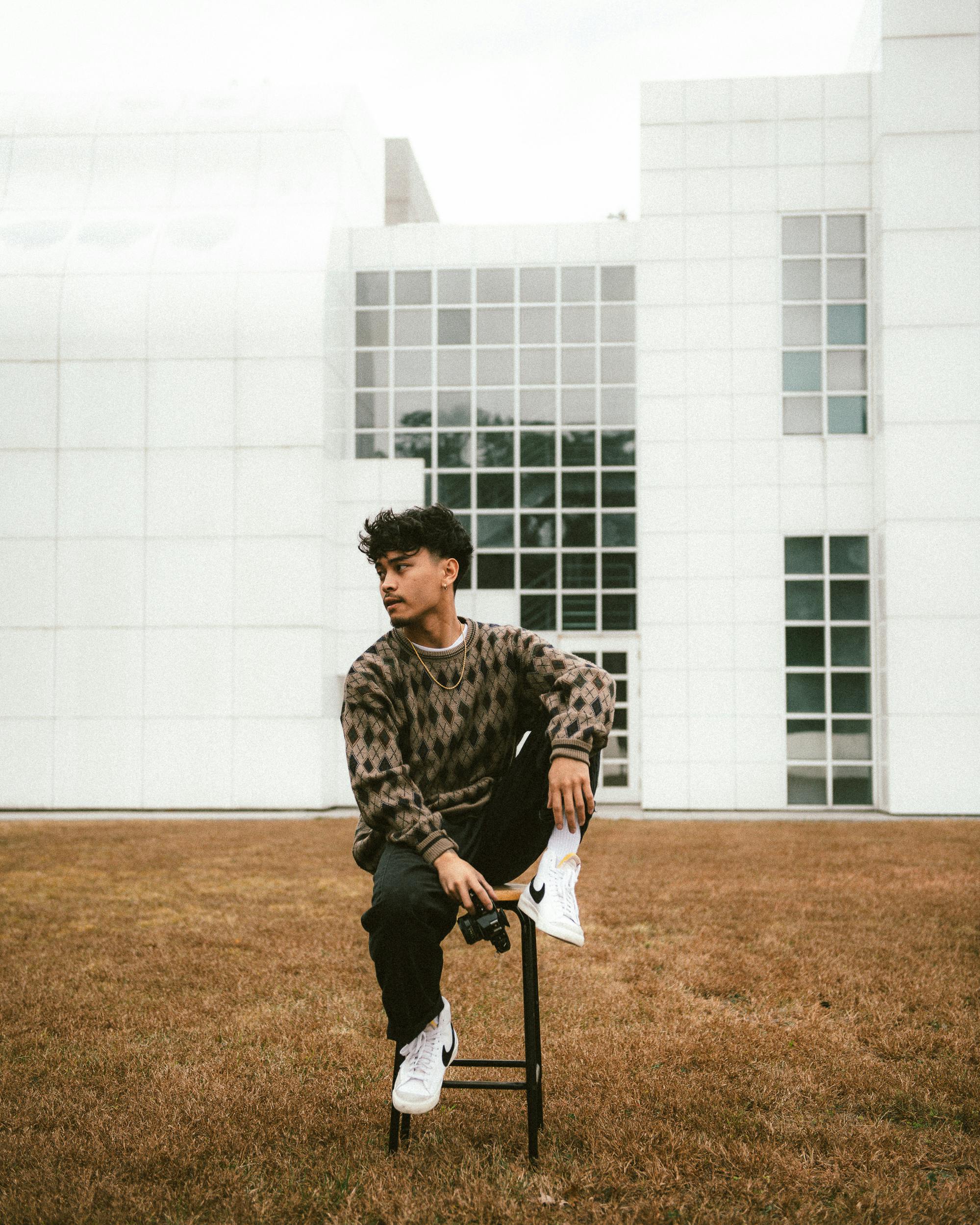 Young man with black hair sitting casually on a stool against a modern building exterior.