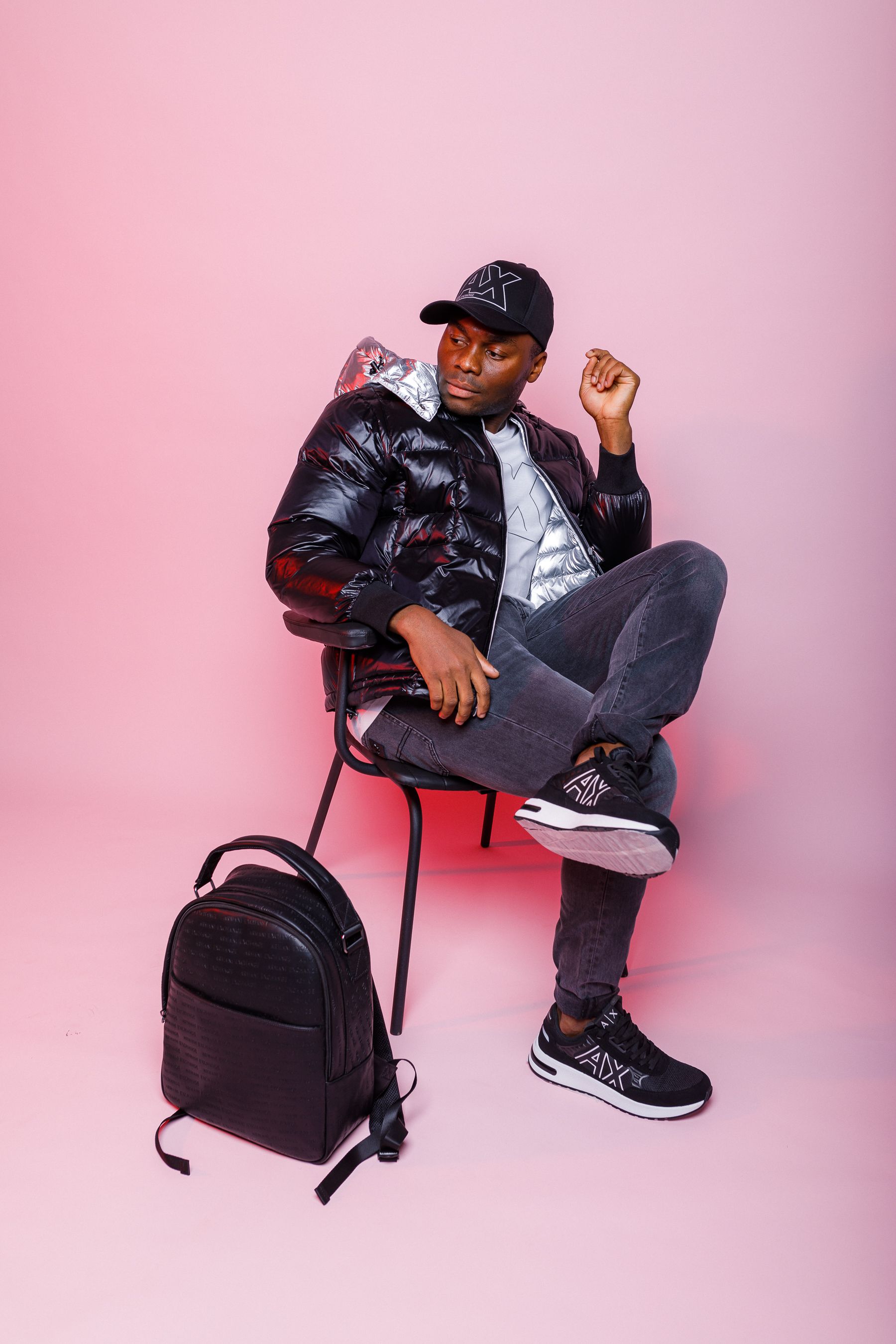 Man sitting on chair with black jacket and backpack on pink background.