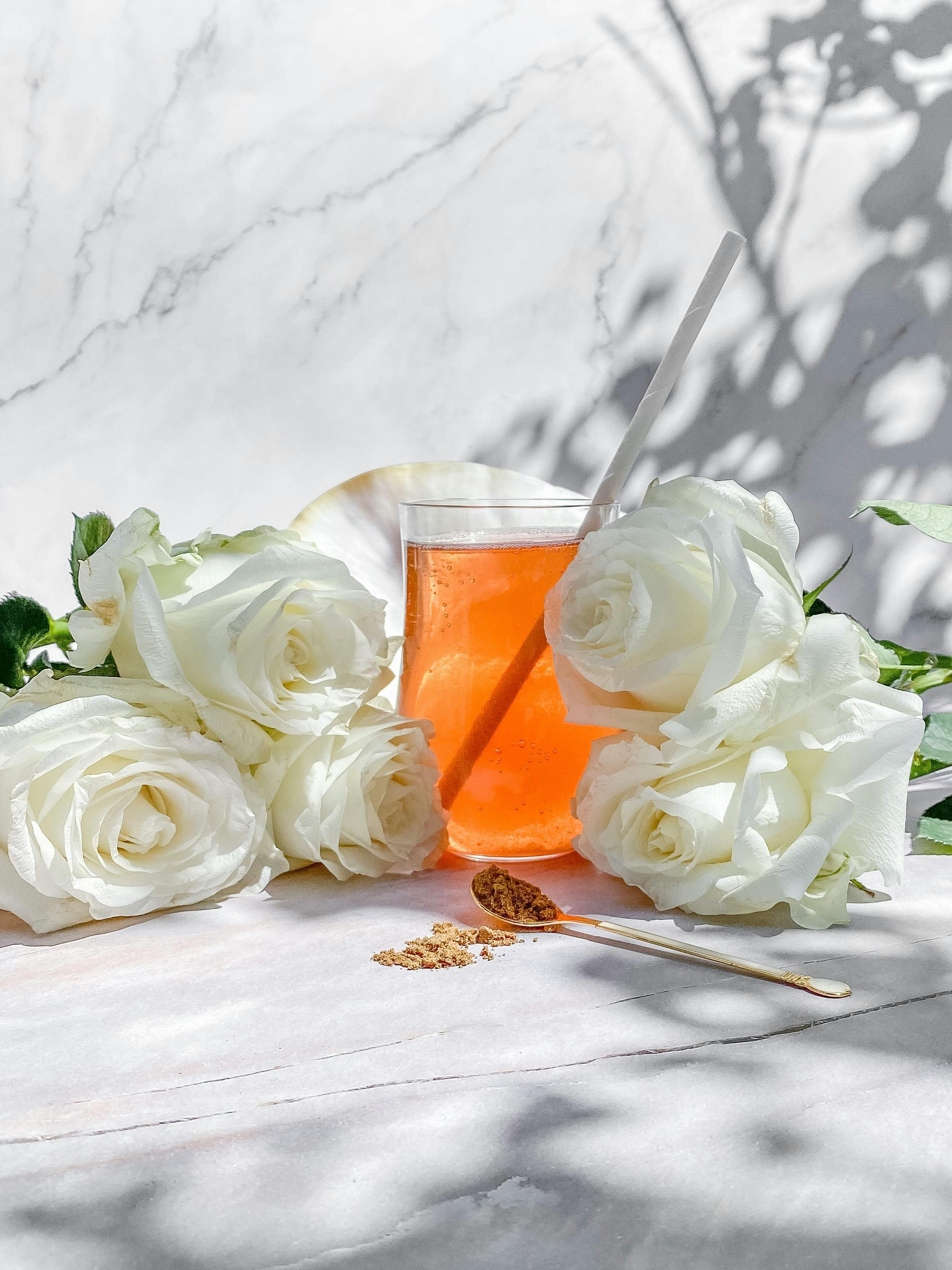Elegant still life of orange drink surrounded by white roses and a spoon on a marble background.