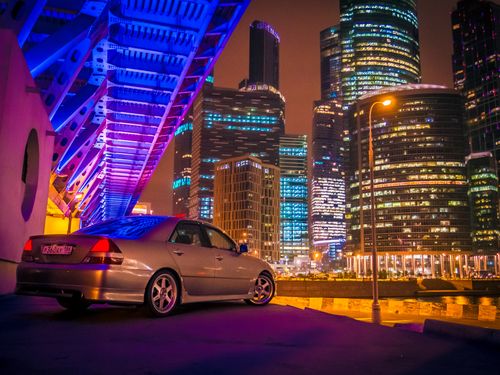 Free A colorful urban scene featuring illuminated skyscrapers and a parked car under a neon-lit bridge. Stock Photo