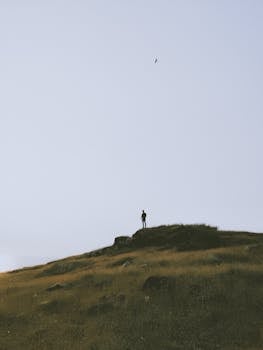 A lone silhouette stands atop a grassy hill at sunrise, conveying tranquility.