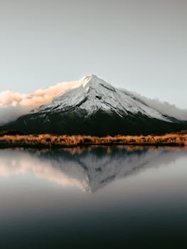 Tranquil view of snow-capped Mount Taranaki reflecting in a serene lake under soft, warm light.