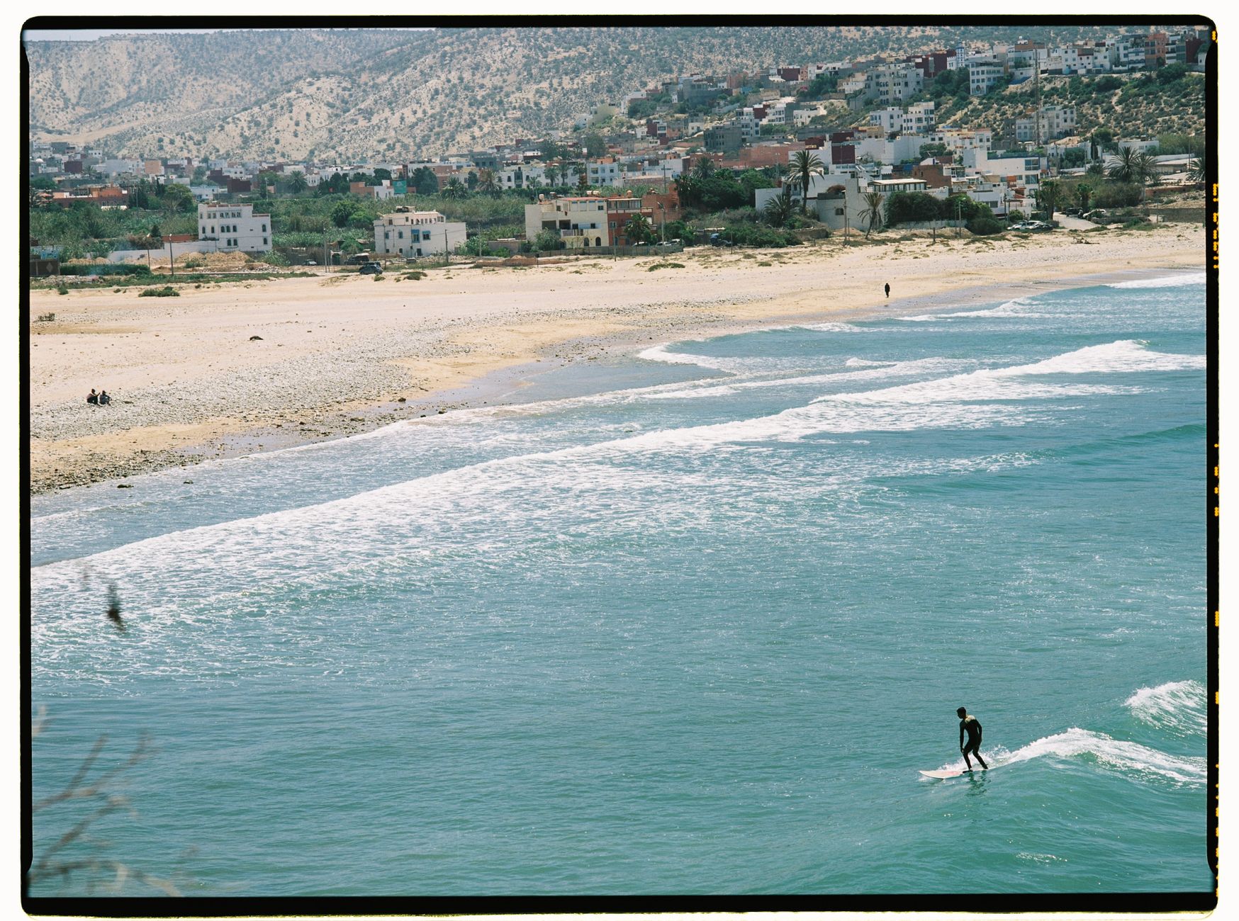 Aerial view of a surfer catching waves along the scenic Moroccan beach with mountains in the background.