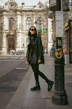 Fashionable woman in sunglasses and leather jacket on a historic city street, with classic architecture in the background.