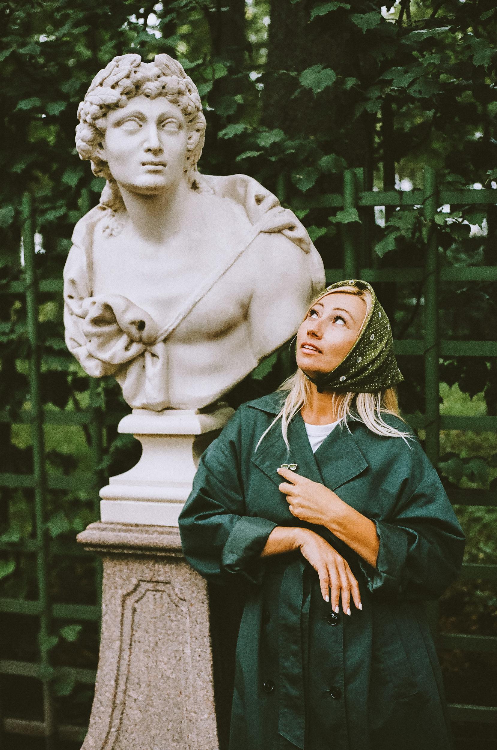 Woman in a green coat with a hair scarf gazing at a classical statue outdoors.