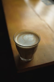 A creamy cappuccino in a glass sits on a sunlit wooden table, perfect for a cozy coffee moment.