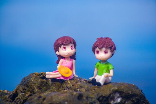 Charming toy figures of a boy and girl sitting on a mossy rock with a blue background, symbolizing youthful innocence.