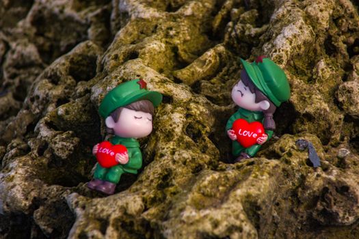 Charming figurines holding love hearts on a textured mossy rock background.