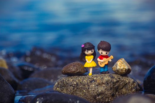 Adorable clay figures of a couple with a guitar on a rocky shore, set against a serene body of water.