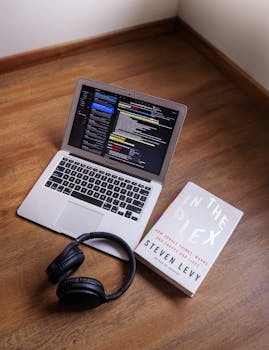 Laptop showing code, 'In the Plex' book, and headphones on wooden floor, captured indoors.