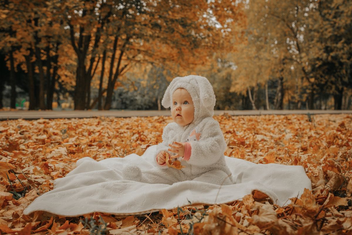 Baby in a cosy white onesie sitting on a blanket surrounded by golden autumn leaves in a park