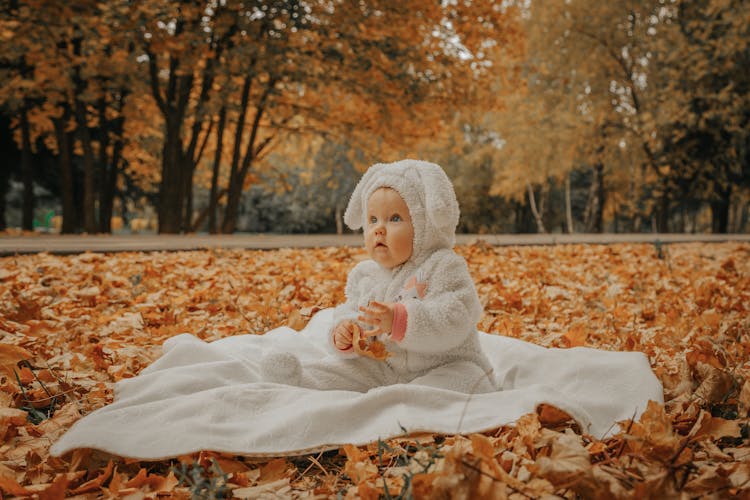 Baby In Bunny Onesie Sitting On Ground Surrounded By Leaves