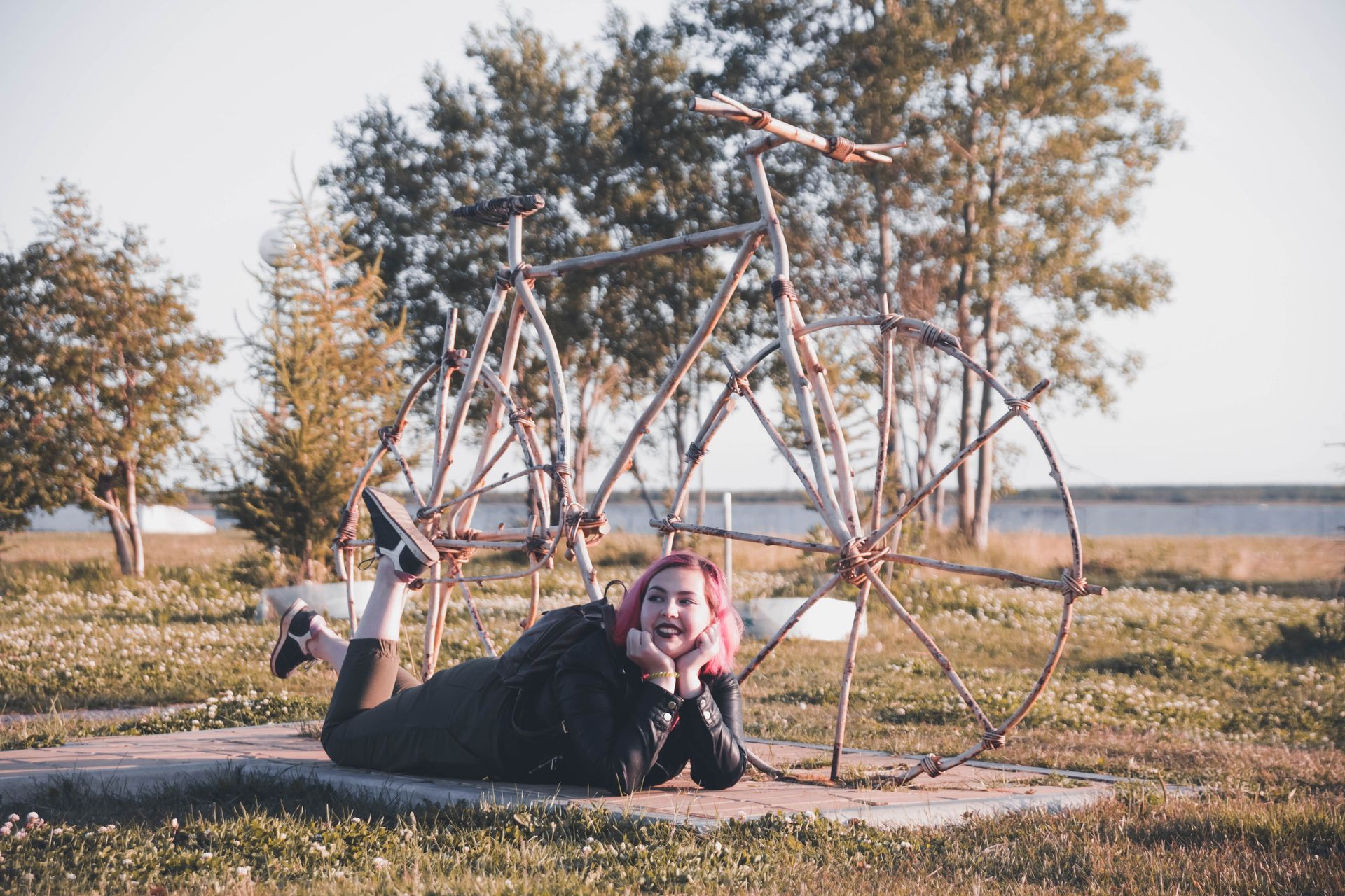 A woman lies on grass next to a large wooden bicycle sculpture in a serene park setting.