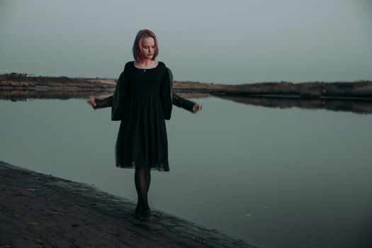 A woman stands contemplatively by a lakeside in a moody twilight setting, dressed in black.