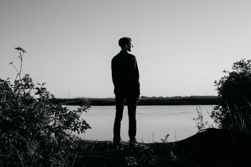 
A Grayscale of a Man Standing in Front of a Lake