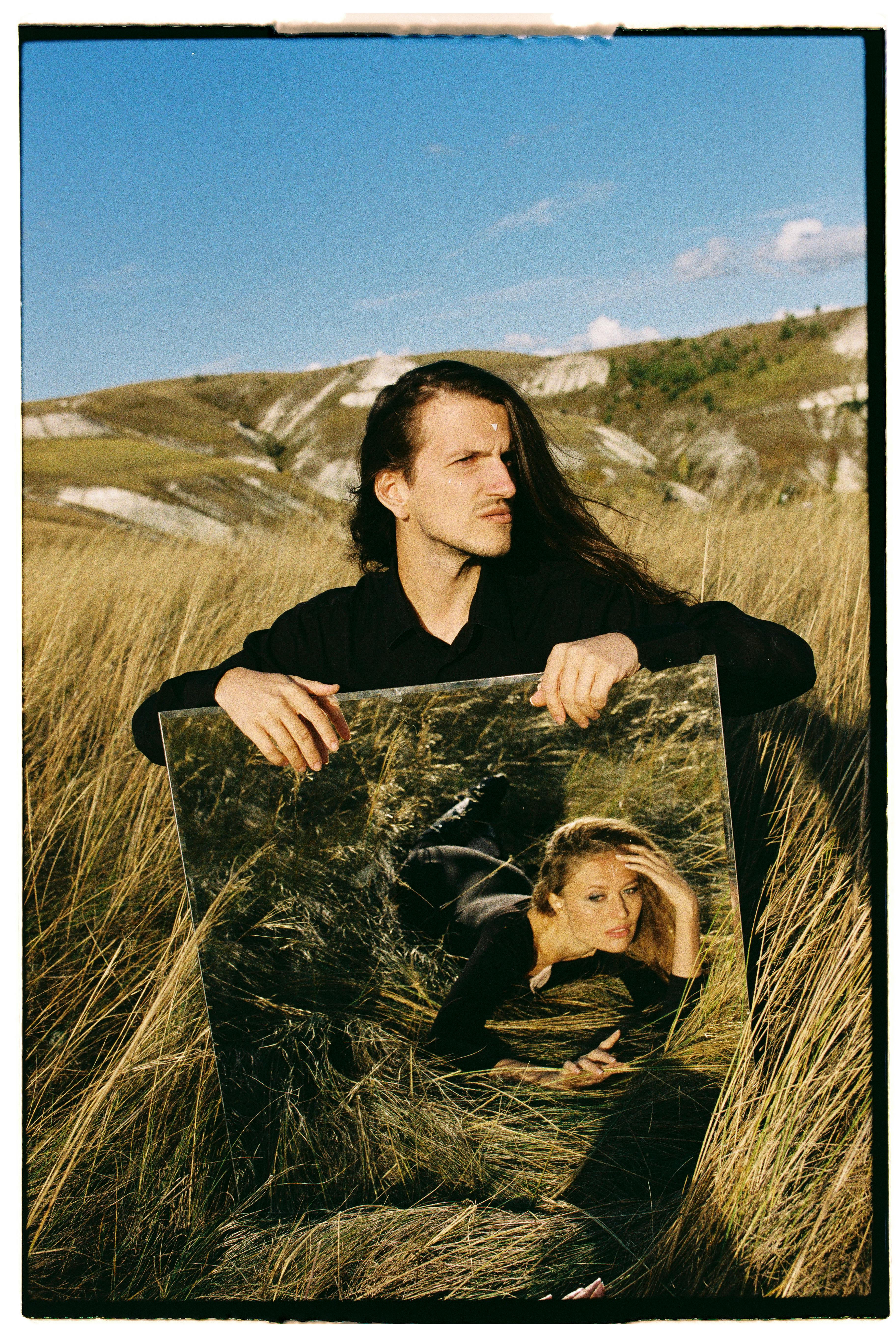 Man holding mirror with artistic reflection in grassy landscape.