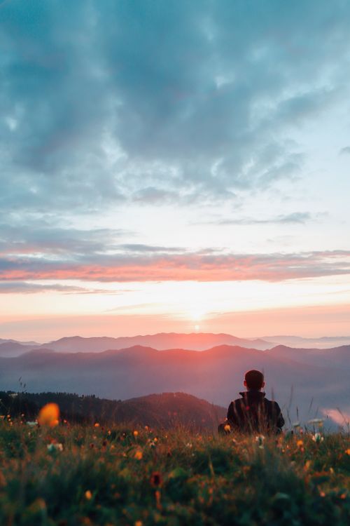 Free Person sits on grass field enjoying a stunning sunset view over misty mountains. Stock Photo