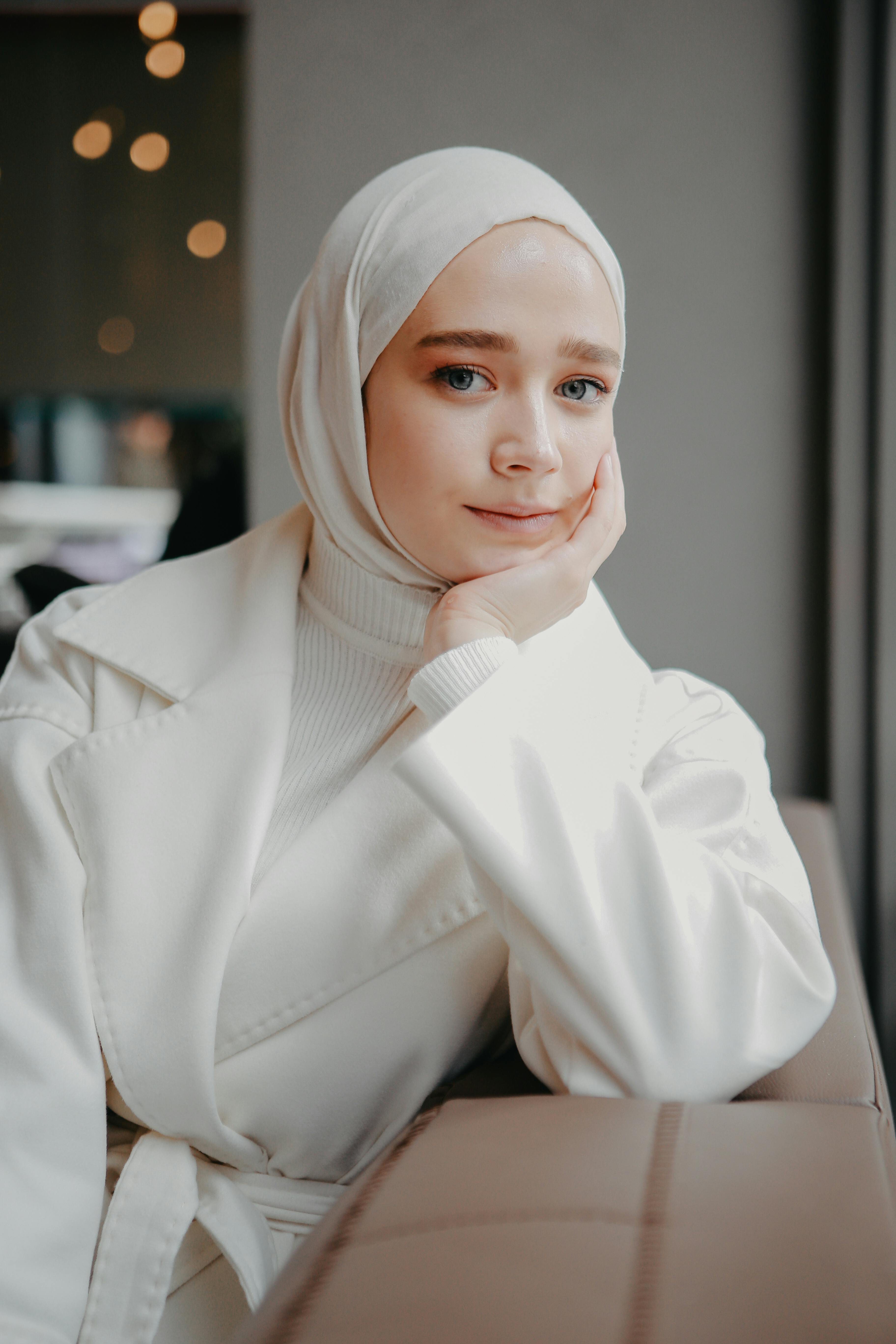 Portrait of a woman in a white hijab and coat, looking thoughtful indoors.