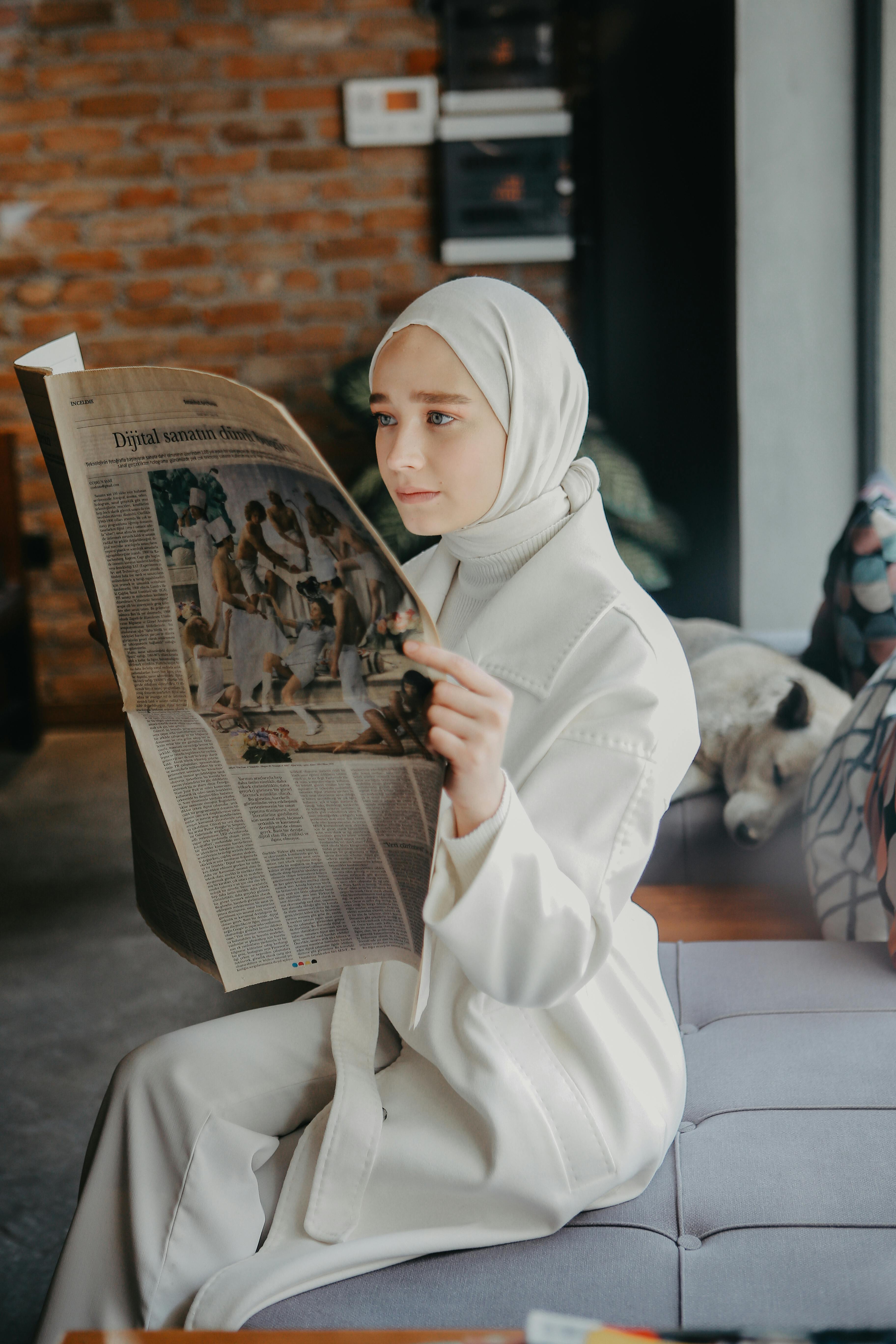 A Muslim woman in modest clothing reads a newspaper while sitting indoors, showcasing a moment of reflection.