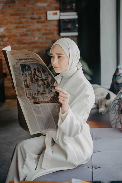 Free A Muslim woman in modest clothing reads a newspaper while sitting indoors, showcasing a moment of reflection. Stock Photo