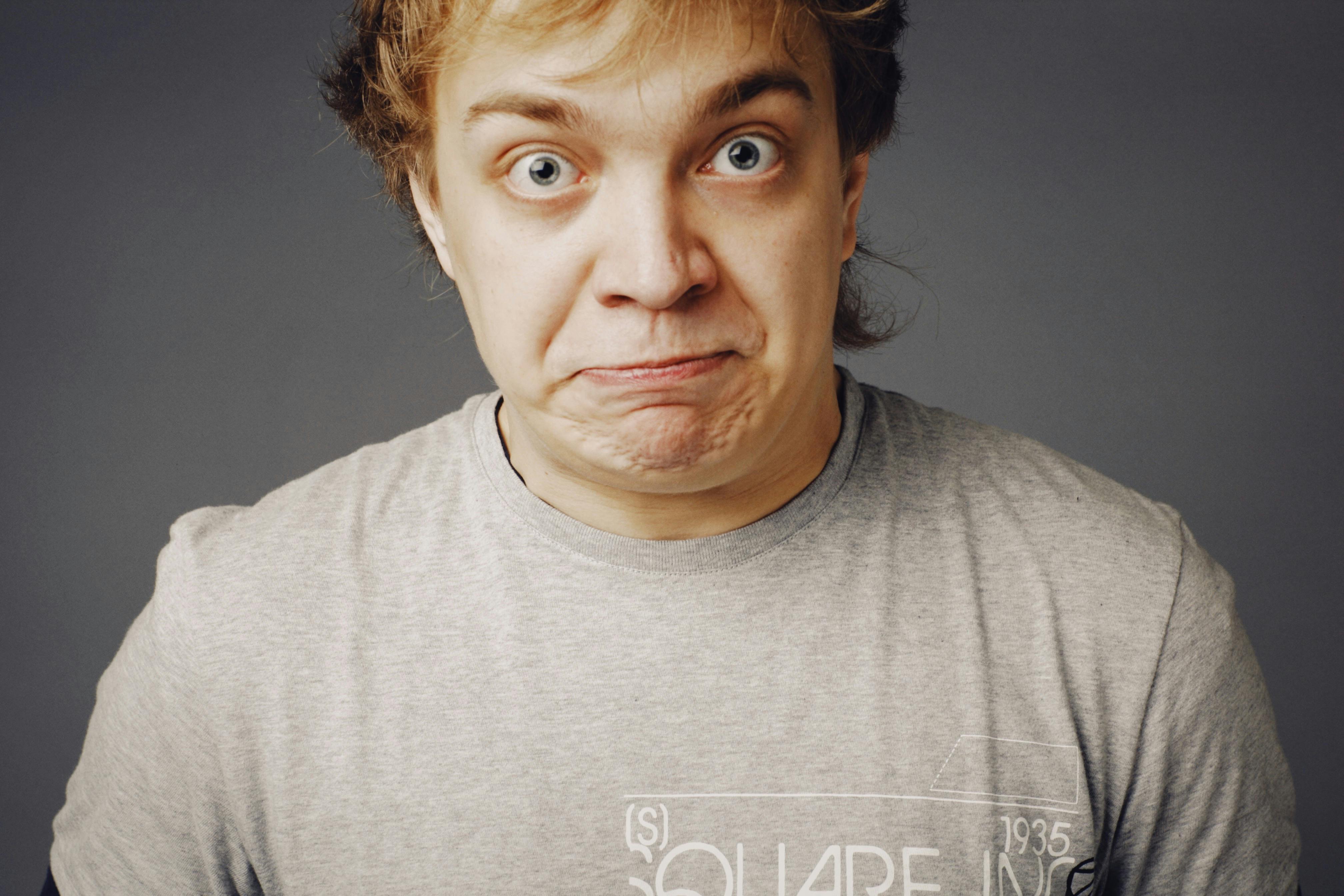 Close-up portrait of a young man in a gray t-shirt making a humorous face, displaying surprise.