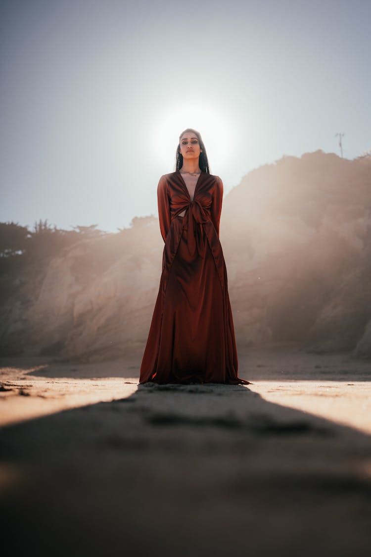 Woman In Long Brown Dress Standing On Beach In Front Of Sun