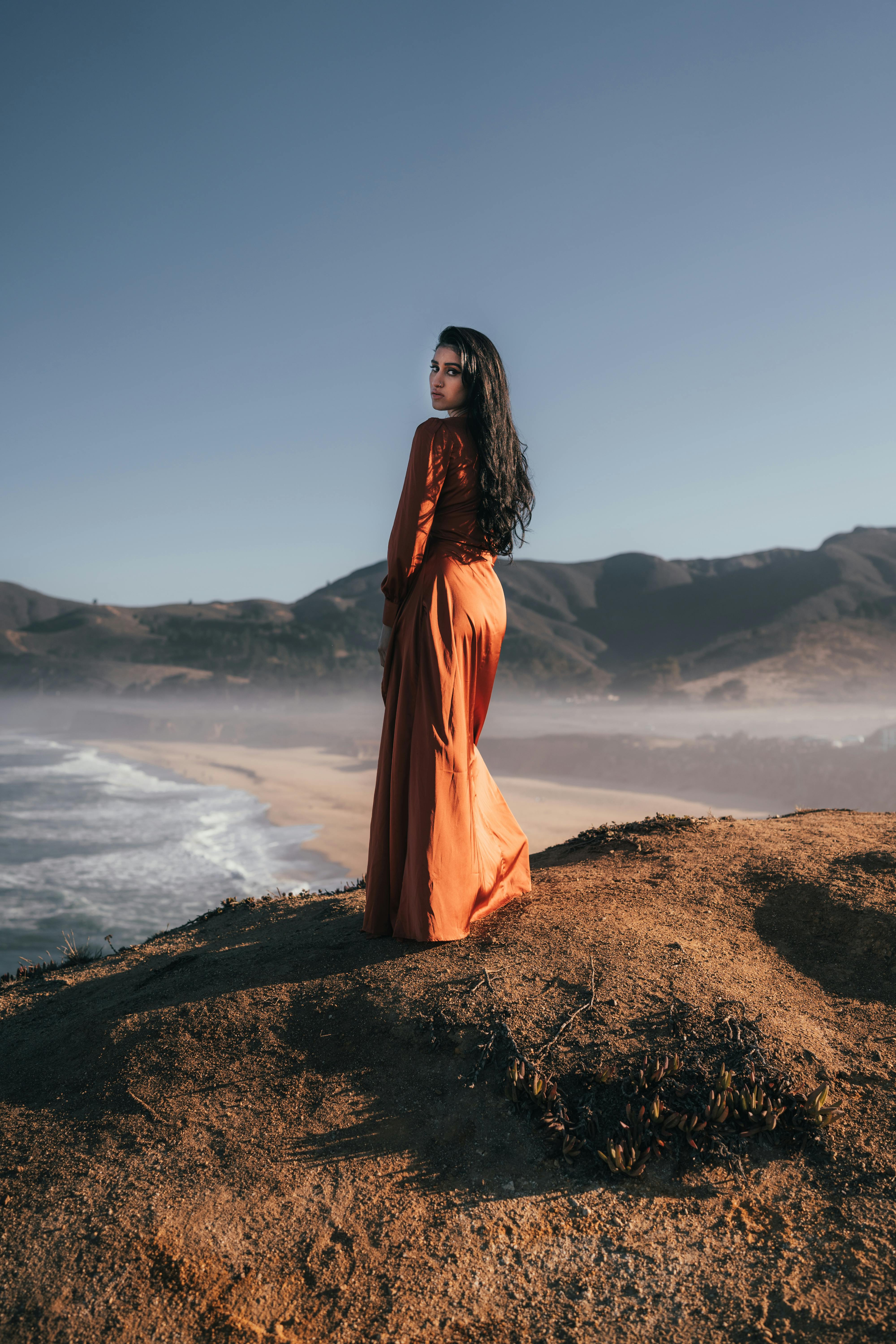 Woman standing on a coastal cliff in a flowing dress, gazing at the ocean under a clear sky.