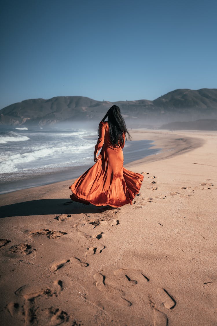 Rear View Of Woman In Long Brown Dress Walking On Beach