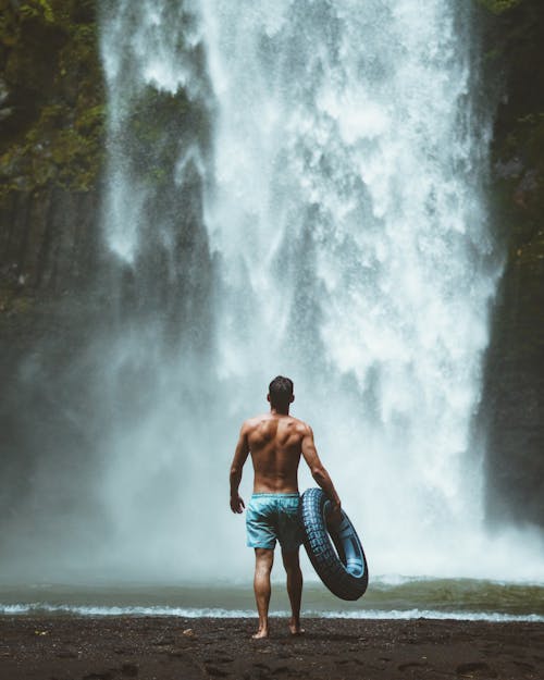Kostenlos Mann, Der Blaue Shorts Hält, Die Fahrzeugreifen Mit Blick Auf Wasserfälle Halten Stock-Foto