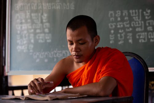 Man in Orange Robe Sitting on Metal Chair Reading a Book