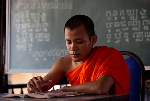 Free A focused Buddhist monk studying at a desk in an educational setting with a chalkboard in the background. Stock Photo